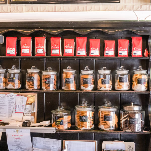 Black café shelving displays red Fazenda coffee bags above glass jars filled with assorted labeled cookies.