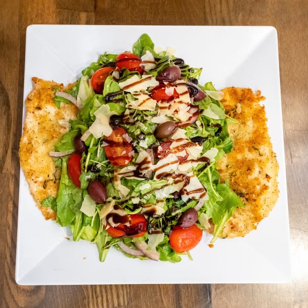Breaded chicken cutlets topped with arugula, tomatoes, olives, shaved Parmesan, and balsamic glaze on a white plate.