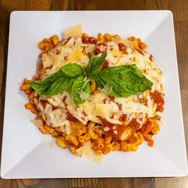 Baked pasta topped with melted mozzarella, tomato sauce, shaved Parmesan, and fresh basil on a white square plate.