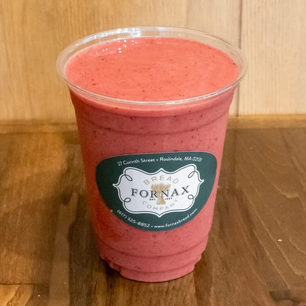 Pink berry smoothie in a clear cup labeled Bread Fornax, resting on a wooden table.