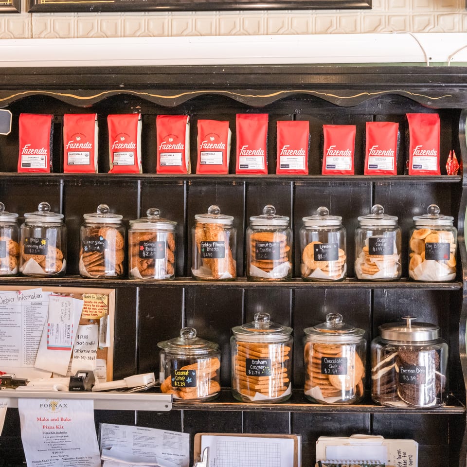 Black wooden café shelving displays red Fazenda coffee bags and labeled glass jars filled with assorted cookies.