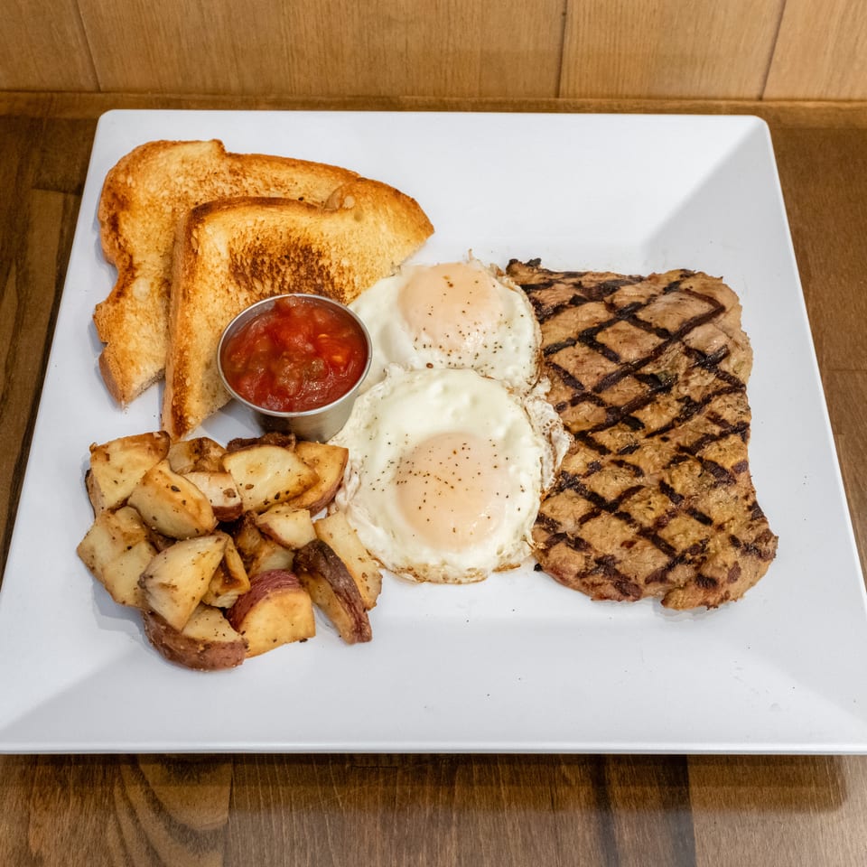 Breakfast platter with grilled steak, two fried eggs, toast, roasted potatoes, and a side of tomato salsa on a white plate.