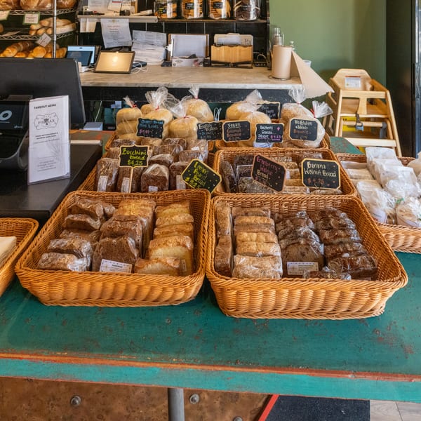 Bakery counter displaying baskets of packaged breads, pastries, labeled specialty loaves, fresh rolls, and a takeaway menu.