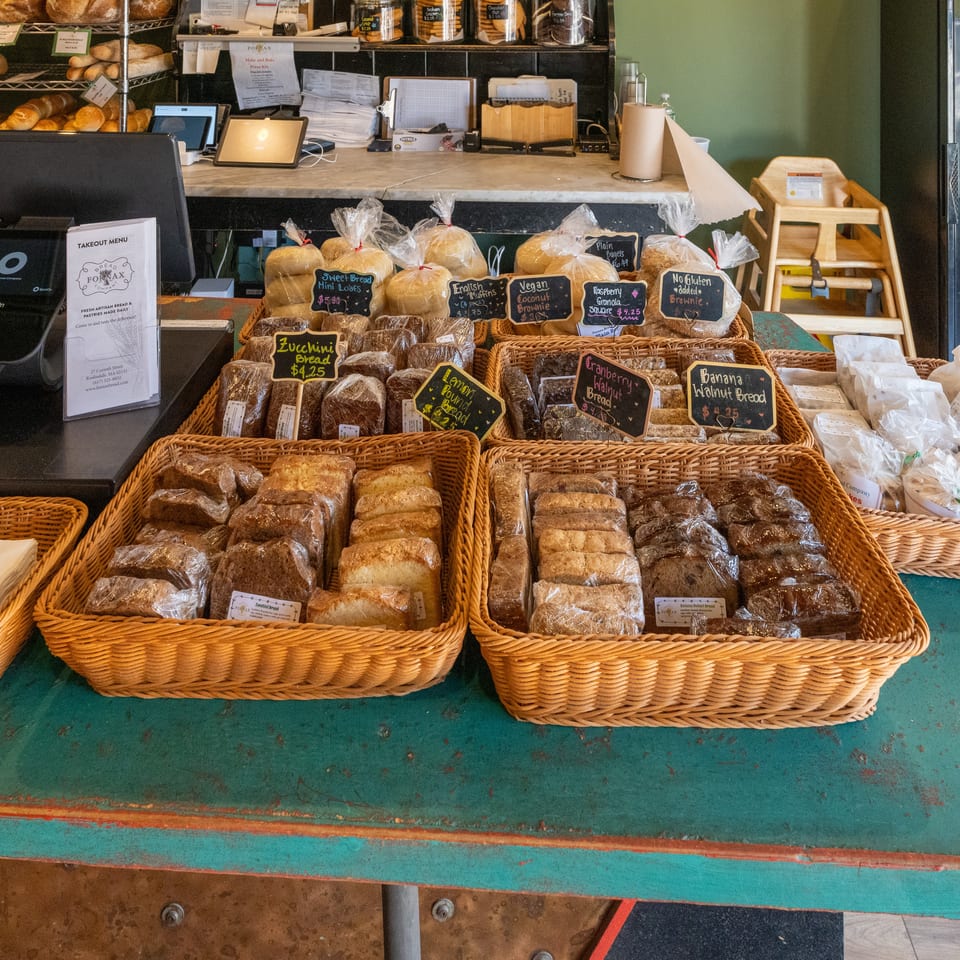 Bakery counter displaying baskets of packaged breads, pastries, labeled specialty loaves, fresh rolls, and a takeaway menu.