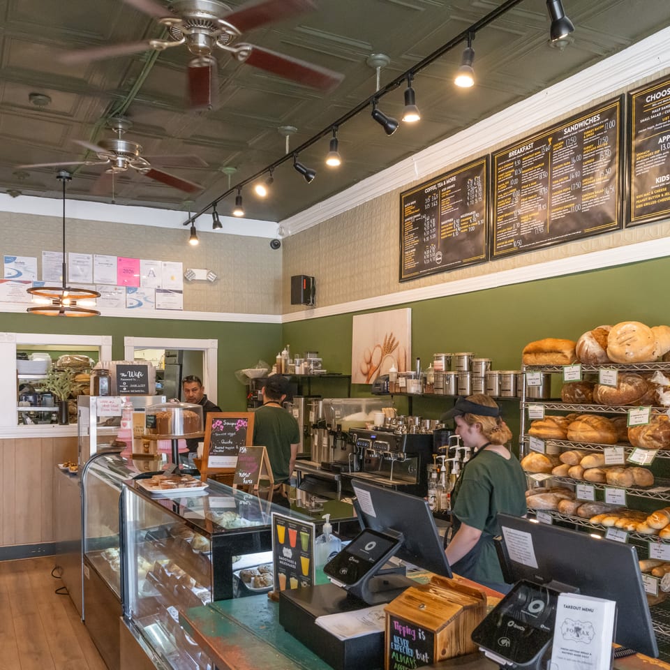 Bakery café counter with staff, pastry displays, espresso equipment, bread racks, and overhead menu boards.